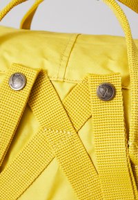 Gul tygryggsäck med korsstygnsremmar, metallaccenter och en texturerad yta. Några framträdande nitar förbättrar designen.