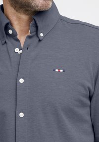 Camicia grigia con bottone, realizzata in un tessuto texturizzato, caratterizzata da un logo ricamato a contrasto in blu, bianco e rosso sul petto, e bottoni bianchi.