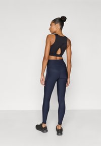 Μαύρο αθλητικό crop top με λεπτομέρεια κλειδαρότρυπας στην πλάτη, συνδυασμένο με ψηλόμεσα σκούρα μπλε κολάν. Το μοντέλο φοράει μαύρα αθλητικά παπούτσια, γυρισμένο προς τα πίσω.