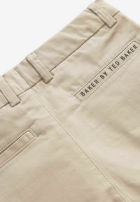 Pantaloni in cotone beige con una superficie liscia, dotati di passanti per cintura e un'etichetta cucita con "BAKER BY TED BAKER" in nero.