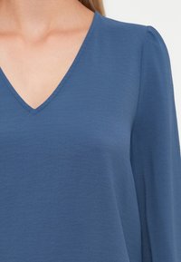 Blå V-hals topp laget av myk, lett stoff. Har lange ermer med subtil puff på skuldrene og en jevn tekstur.