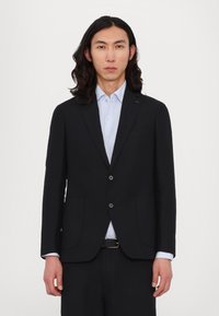 Schwarzer Blazer mit zwei Vorder- taschen, Ein-Knopf-Verschluss, kombiniert mit einem hellblauen Hemd. Auffällige glatte Textur und taillierte Passform.