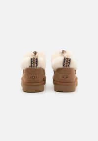 Zapatillas de ante marrón con un suave ribete de color crema, intrincados acentos tejidos y una suela de goma flexible. Marca UGG visible en la parte posterior.