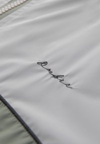 Lys grå stof med en glat tekstur, prydet med delikat sort broderi af ordet "emilie." Indeholder kontrastfarvede grønne detaljer langs kanterne.