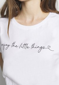 Vit bomulls t-shirt med svart kursivtryck som säger "njut av de små sakerna" med en liten hjärtaccent. Rund halsringning.