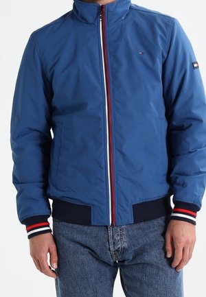 Niebieska kurtka na suwak z czerwono-biało-granatowymi pasiastymi wykończeniami na mankietach i pasie, noszona przez osobę z rękami swobodnie opuszczonymi wzdłuż ciała.