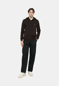 Felpa nera con zip e cappuccio con coulisse, con logo sul petto, abbinata a pantaloni neri a taglio rilassato e scarpe da ginnastica di colore chiaro.