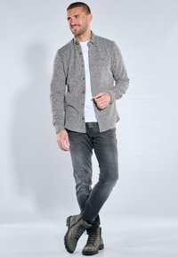 Grau strukturiertes Hemd zum Knöpfen über einem weißen T-Shirt, kombiniert mit dunklen Jeans und olivgrünen Stiefeln. Einfache Hardware-Details sichtbar.