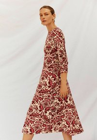 Bloemenpatroond jurk in rood en crèmekleur, halflang met driekwart mouwen, met een getailleerde bovenkant die uitloopt bij de taille.