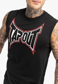 Juodas medvilnės be rankovių marškinėliai su ryškiu raudonu ir baltu "TAPOUT" logotipu priekyje. Lygus audinys, prigludusi forma ir apvalus iškirpimas.