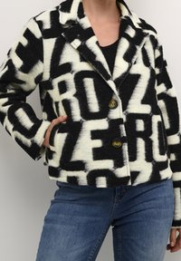Svart och krämfärgad mönstrad jacka med ordet 'ZERO' genomgående. Tillverkad av texturerad tyg, med kavajkrage och två knappar.