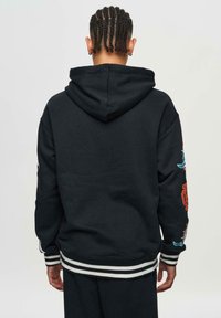 Sweatshirt à capuche noire avec des poignets et un ourlet à rayures blanches. Présente des graphismes colorés sur les manches. Tissu doux avec une coupe décontractée.