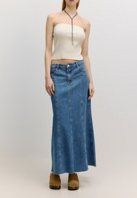 Trägersloses, geripptes, hellbeiges Top, kombiniert mit einem langen blauen Denimrock mit vertikalen Nähten und fransigem Saum. Braune, hochhackige Clogs runden den Look ab.