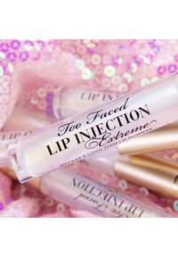 Doorzichtig lipplumper met een metallic dop, voorzien van een zwart label in scriptstijl. De achtergrond is bedekt met roze pailletten, wat een textuurcontrast creëert.