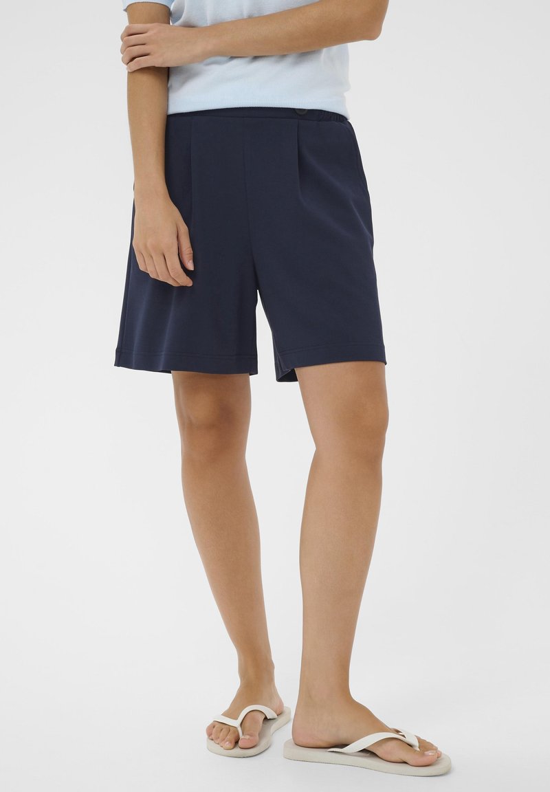 Personne portant un short bleu marine jusqu'aux genoux, un haut bleu clair et des tongs blanches, debout sur un fond blanc.