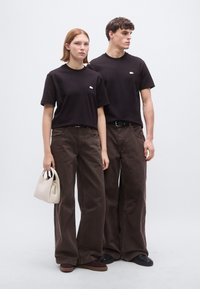 Μαύρα T-shirt, φαρδιά καφέ παντελόνια και μια ελαφριά τσάντα χειρός. Τα T-shirt έχουν ένα μικρό λογότυπο· τα παντελόνια έχουν φαρδιά γραμμή στα πόδια. Τα παπούτσια είναι σκούρα.