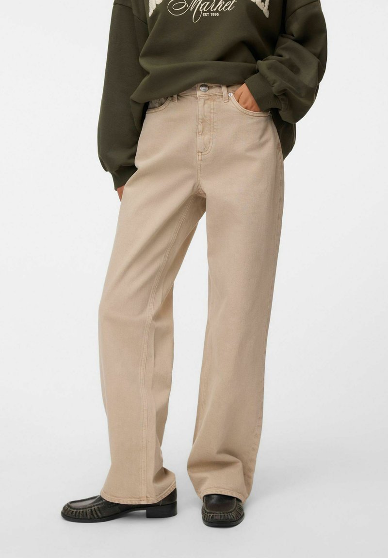 Personne portant un pantalon beige à jambes larges, un sweat-shirt vert foncé et des mocassins noirs, les mains dans les poches, debout devant un fond uni.