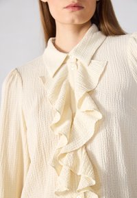 Blusa crema con detalle fruncido en el frente, tejido texturizado, cuello puntiagudo y pequeños botones. Presenta mangas largas y sutiles rayas verticales.