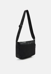 Svart axelväska med en slät, glansig frampanel och matt tygkropp. Har en justerbar rem och ett fack med dragkedja.
