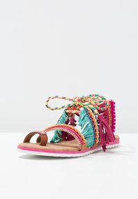 Sandale colorée avec une frange multicolore et des accents de perles. La semelle est rose, avec un design plat et une boucle pour les orteils pour un soutien.