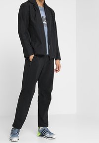 Svart lättviktsjacka med huva, tillsammans med mörka byxor och grå sportskor med gröna detaljer. Strukturerad tyg genom hela plagget.