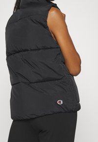 Black puffer vest with a high collar, padded design, armholes, and a small logo in red and white on the lower left side. Textured finish.