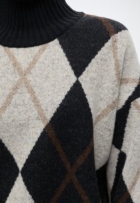 Pull en tricot noir, beige et marron à motif losanges avec un col roulé côtelé noir.