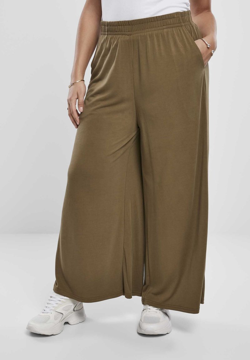 Personne portant un pantalon ample et large de couleur vert olive avec une taille élastique et des baskets blanches, debout avec une main dans la poche.