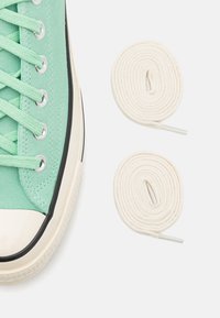 Mátově zelená plátěná teniska s bílou gumovou špičkou a černým lemováním. Obsahuje dva namotané krémově zbarvené tkaničky s plastovými konci.