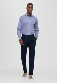 Camicia a righe blu e bianche a maniche lunghe con colletto, abbinata a pantaloni scuri e sneakers marrone chiaro. Vestibilità pulita e sartoriale.