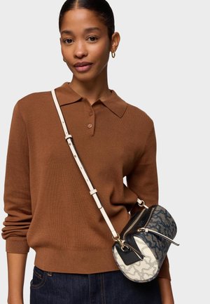 Femme aux cheveux foncés attachés en un chignon bas, portant des boucles d'oreilles créoles dorées, un pull marron boutonné, un jean foncé et un sac bandoulière à motif avec une sangle blanche.