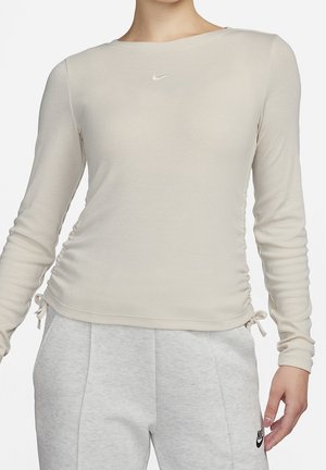 Vrouw draagt een beige geribd shirt met lange mouwen en zijknopen, gecombineerd met lichtgrijze Nike joggingbroek.