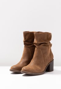 Bottines en daim marron avec tige souple, bout arrondi et talon empilé, présentant une finition texturée et des détails de couture.