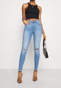 Jean skinny en denim bleu clair avec des genoux déchirés, taille haute, présentant un design de poche. Associé à un crop top noir et des talons avec lanières.