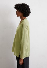 Maglione verde chiaro, morbido e svasato con scollatura a V, maniche lunghe e spacchi laterali, che mette in risalto un tessuto morbido e testurizzato.