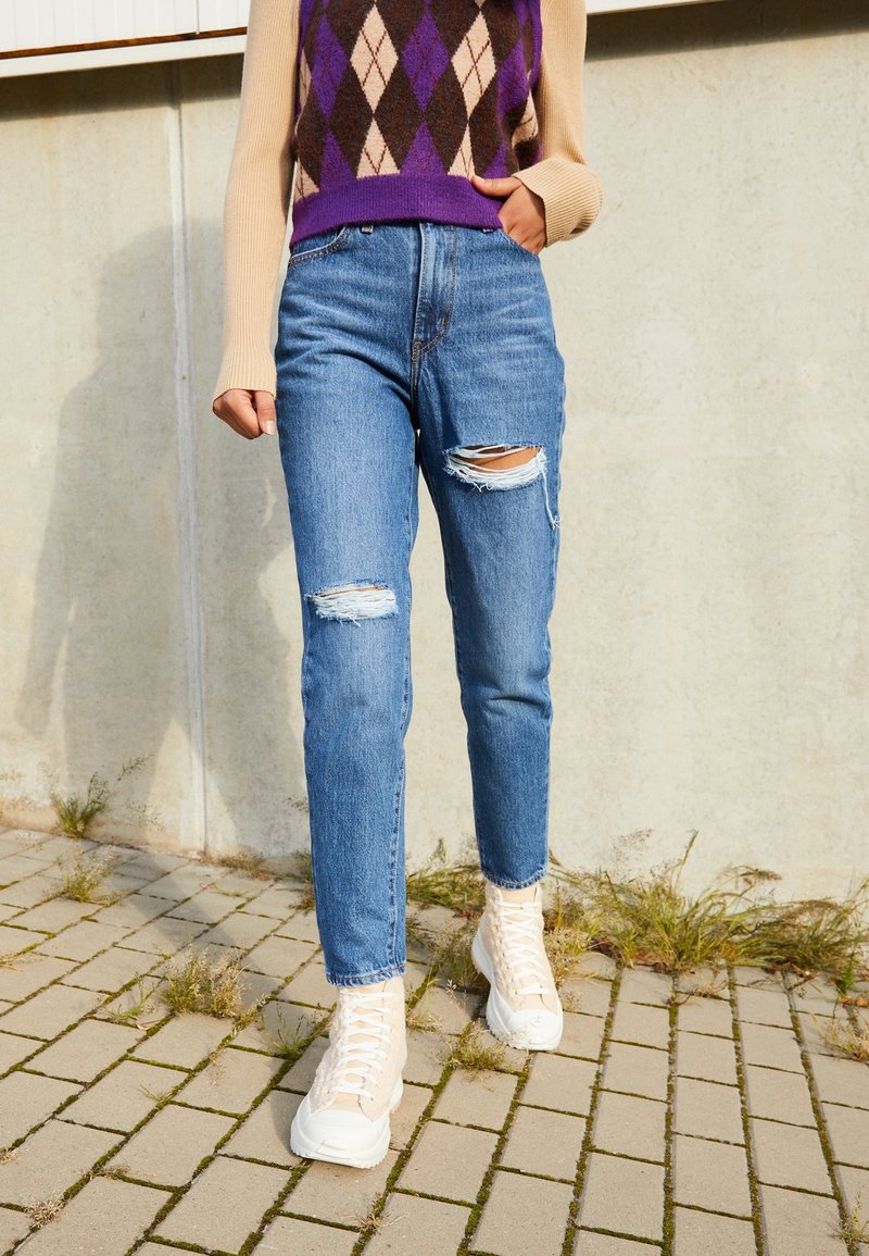 Personne portant un jean bleu déchiré, des baskets montantes beiges et un pull losangé violet et beige debout sur un sol pavé.