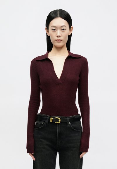 Γυναίκα με ίσια μαύρα μαλλιά που φοράει μακρυμάνικη μπορντό μπλούζα με βαθύ V και μαύρα τζιν με μαύρη ζώνη με χρυσή αγκράφα.