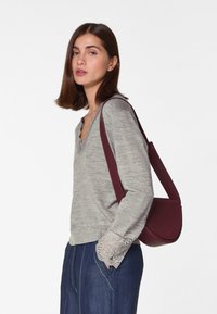 Graues Langarmshirt mit gewelltem Ausschnitt und Spitzenakzenten, kombiniert mit einer burgunderroten Umhängetasche, getragen mit dunkelblauen Jeanshosen.