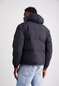 Doudoune noire avec capuche, dotée d'un design matelassé et de poignets élastiques. Portée avec un jean en denim bleu clair.