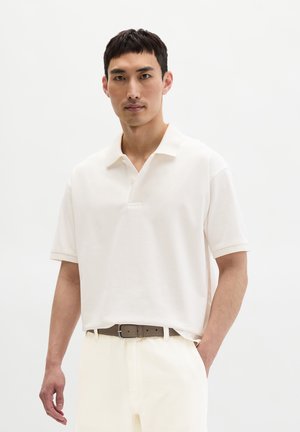 Man draagt een off-white polo met korte mouwen, ingestopt in lichtgekleurde broek met een bruine riem, staand tegen een effen witte achtergrond.