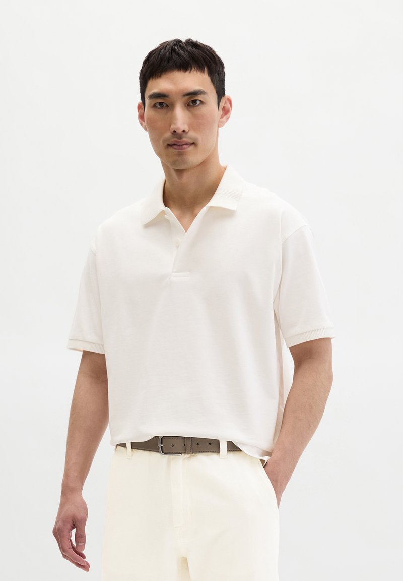 Uomo che indossa una polo a maniche corte color avorio infilata in pantaloni chiari con cintura marrone, in piedi davanti a uno sfondo bianco uniforme.