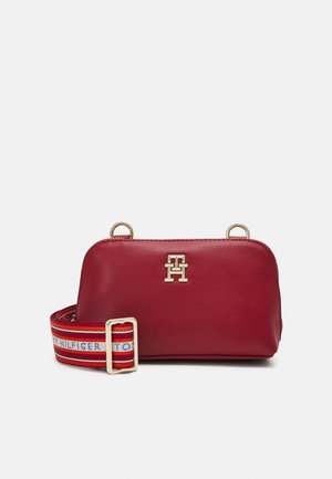 Bolso bandolera de cuero rojo con logo "TH" en tono dorado y correa ajustable de tela de marca roja, blanca y azul sobre un fondo gris.