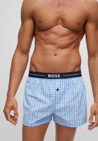 Boxers à carreaux bleus et blancs avec une taille noire portant le nom de la marque "BOSS" en blanc. Détail de bouton à l'avant.