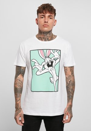 Homme avec des tatouages portant un t-shirt blanc arborant un grand dessin animé de Bugs Bunny dans un carré vert, debout sur un fond uni.