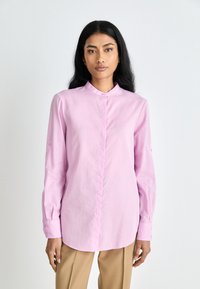 Camicia a maniche lunghe rosa chiaro con colletto alla cinese. Presenta una chiusura frontale con bottoni e maniche arrotolate con dettagli in bottone. Tessuto morbido e liscio.