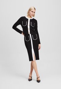 Black knit dress set with white trim, featuring cropped jacket with gold buttons and four pockets, and a skirt with a front slit and pockets.