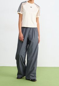 Krämfärgad kortärmad skjorta med svarta meshdetaljer och logotyp, tillsammans med löst sittande mörkgrå byxor med elastisk midja.