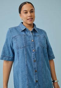 Femme aux cheveux foncés portant une chemise en jean bleu à manches courtes boutonnée, debout devant un fond bleu clair.