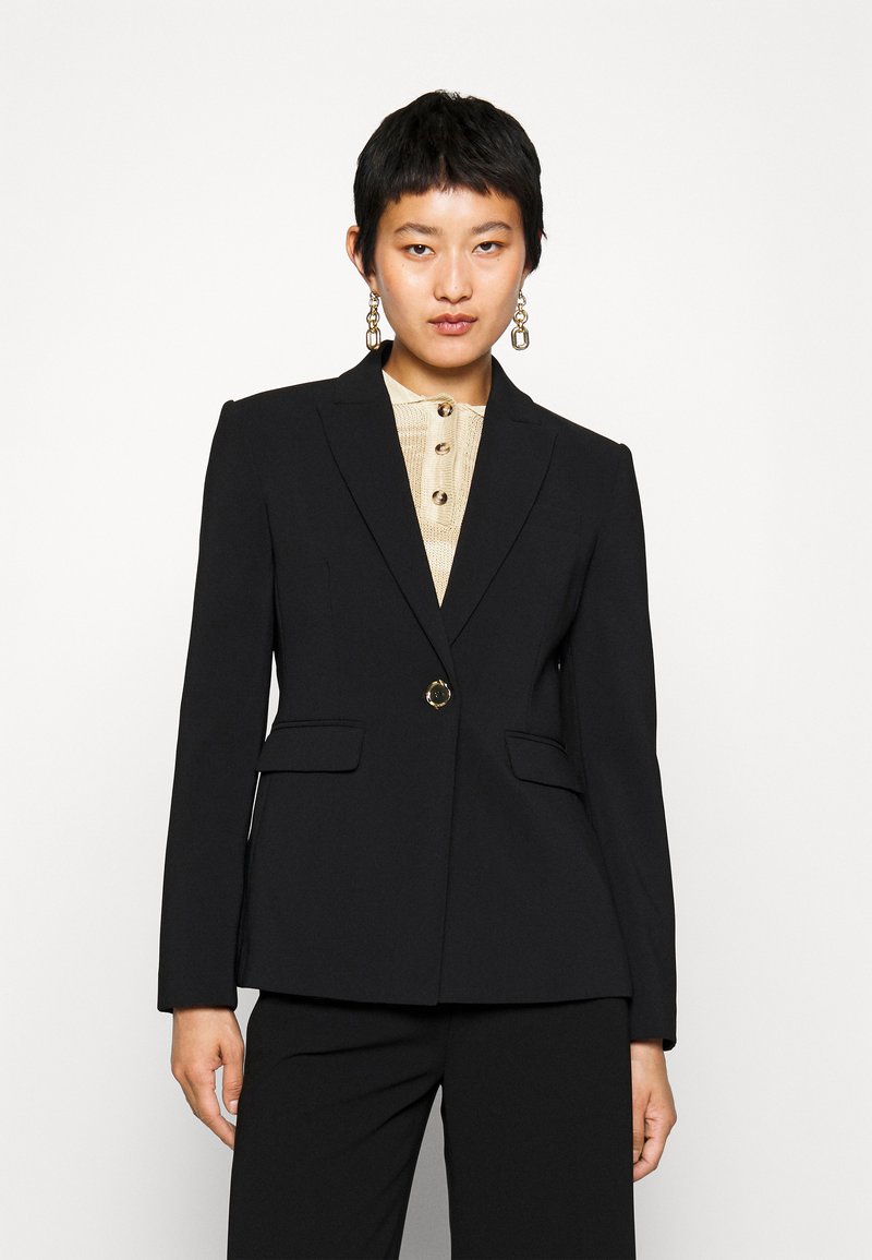 Blazer nero su misura con rever a ragno, chiusura con un solo bottone dorato e due tasche frontali. Indossato sopra una camicia beige con texture.
