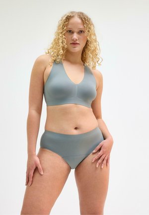 Kvinde med krøllet blondt hår iført et matchende lysegråt sømløst BH- og undertøjsæt, stående mod en ensfarvet hvid baggrund.